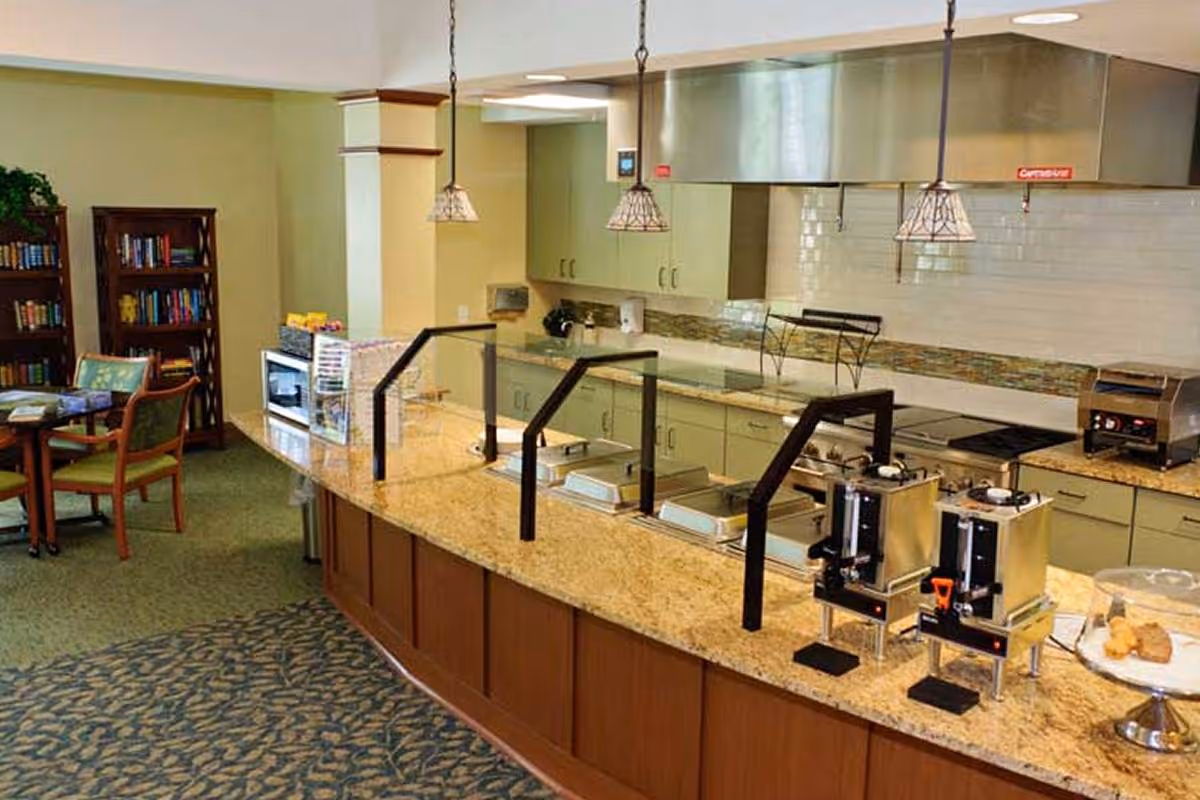 A senior living facility kitchen area with a long granite countertop featuring food warmers and beverage dispensers. Behind the counter are cabinets, a stove, and a ventilation hood. To the left, there is a small seating area with a table, chairs, and bookshelves filled with books.
