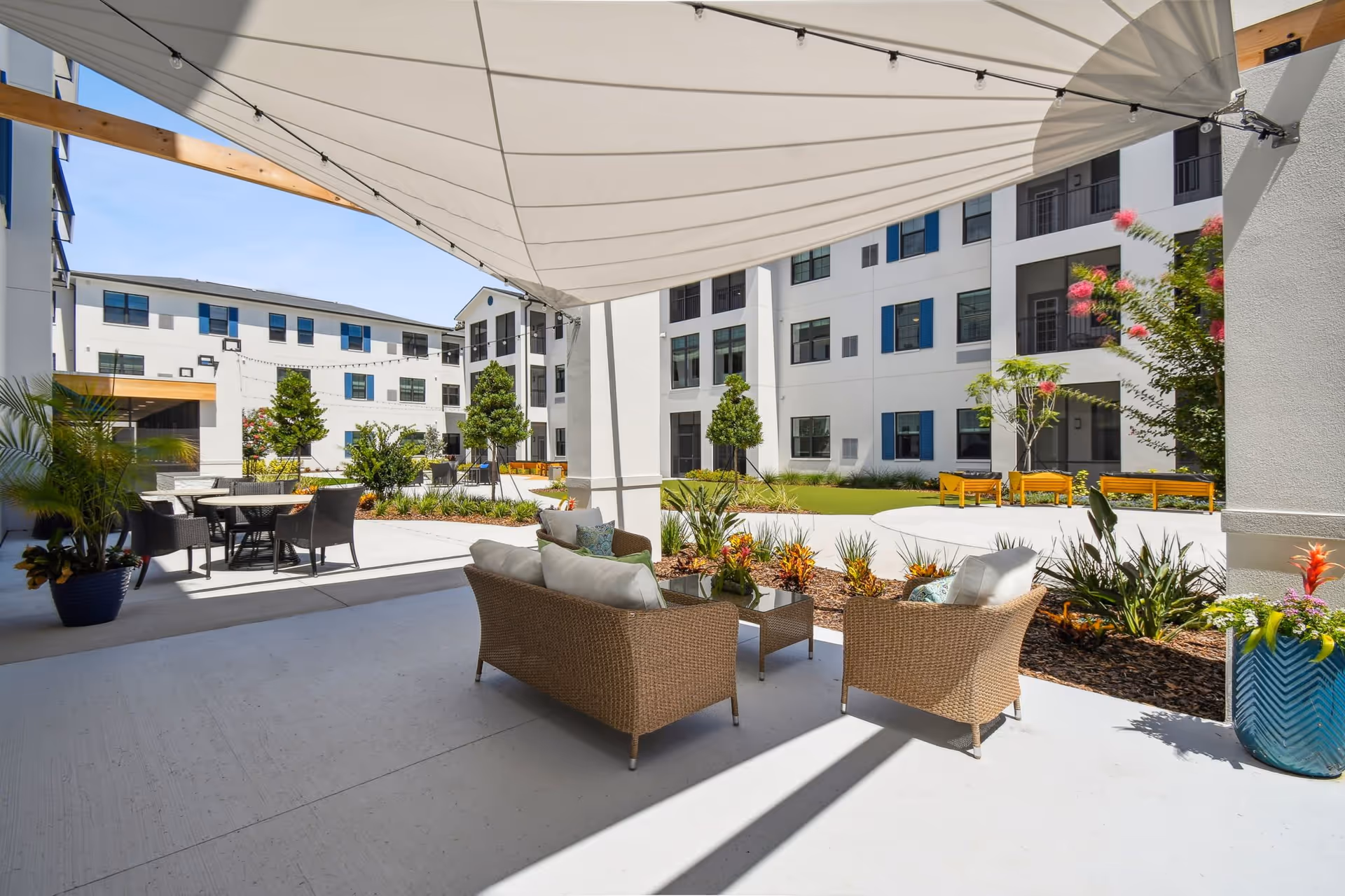 Outdoor seating area at Aden Senior Living with wicker furniture including a sofa and chairs under a large white canopy. The area overlooks a courtyard with landscaped plants, trees, benches, and a multi-story white building with blue window shutters in the background.