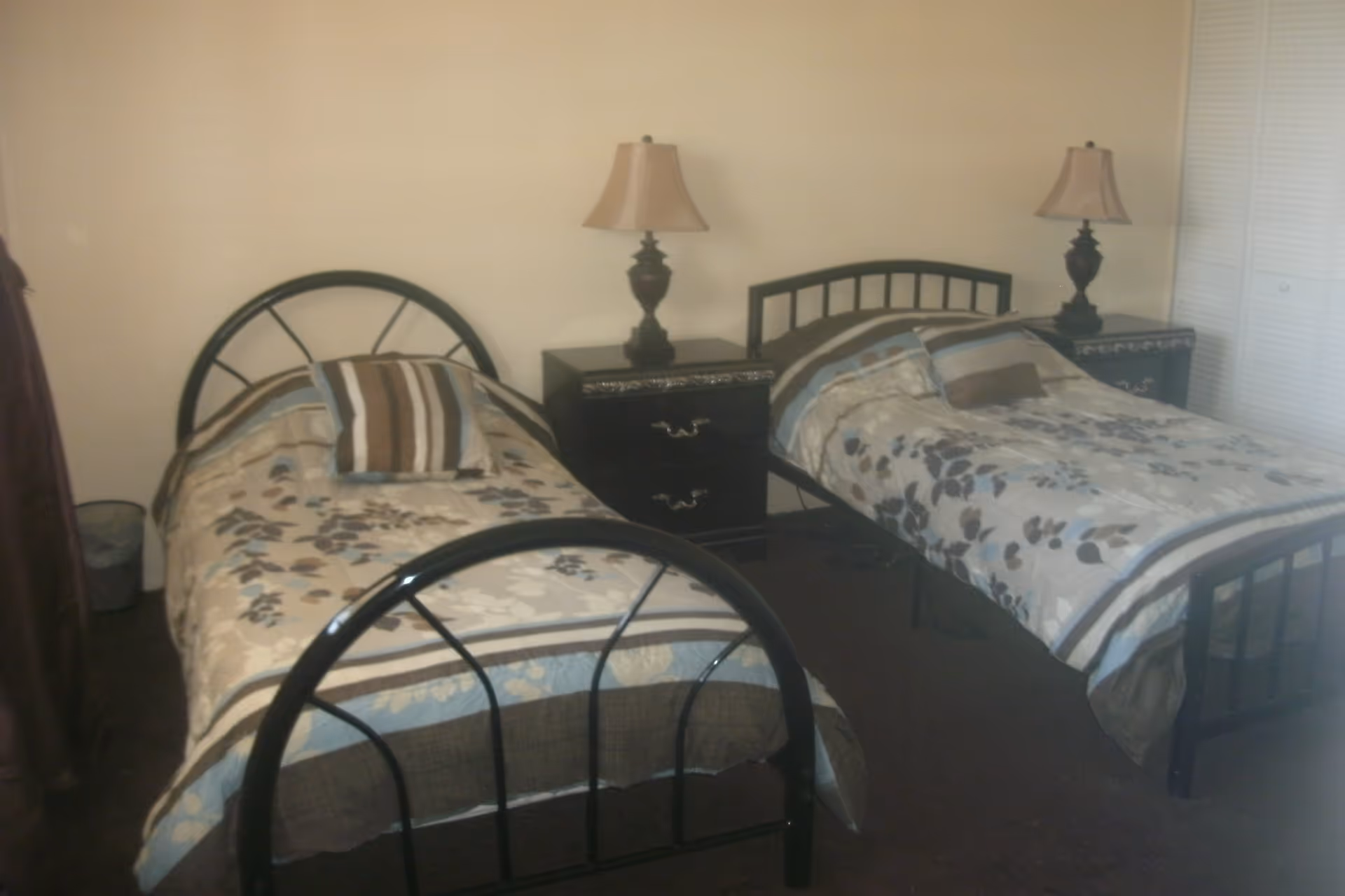 A bedroom with two single beds, each with a floral patterned bedspread and a striped pillow. Between the beds is a black nightstand with two drawers and a lamp on top. The walls are plain and light-colored, and there is a closet with white louvered doors on the right side.