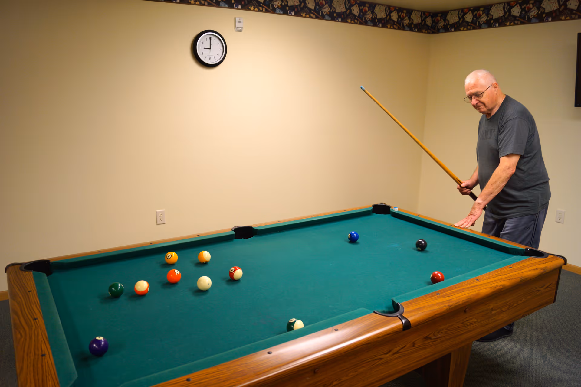 An elderly man playing pool in a room with beige walls and a clock on the wall showing 10:10. The pool table has several balls scattered on the green felt surface.