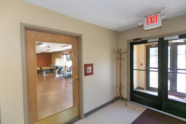 Interior view of a hallway in a nursing and rehabilitation center showing a wooden door with a large glass panel leading to a room with exercise equipment. To the right, there is a double glass exit door with an illuminated exit sign above it and a wooden coat rack beside the door.