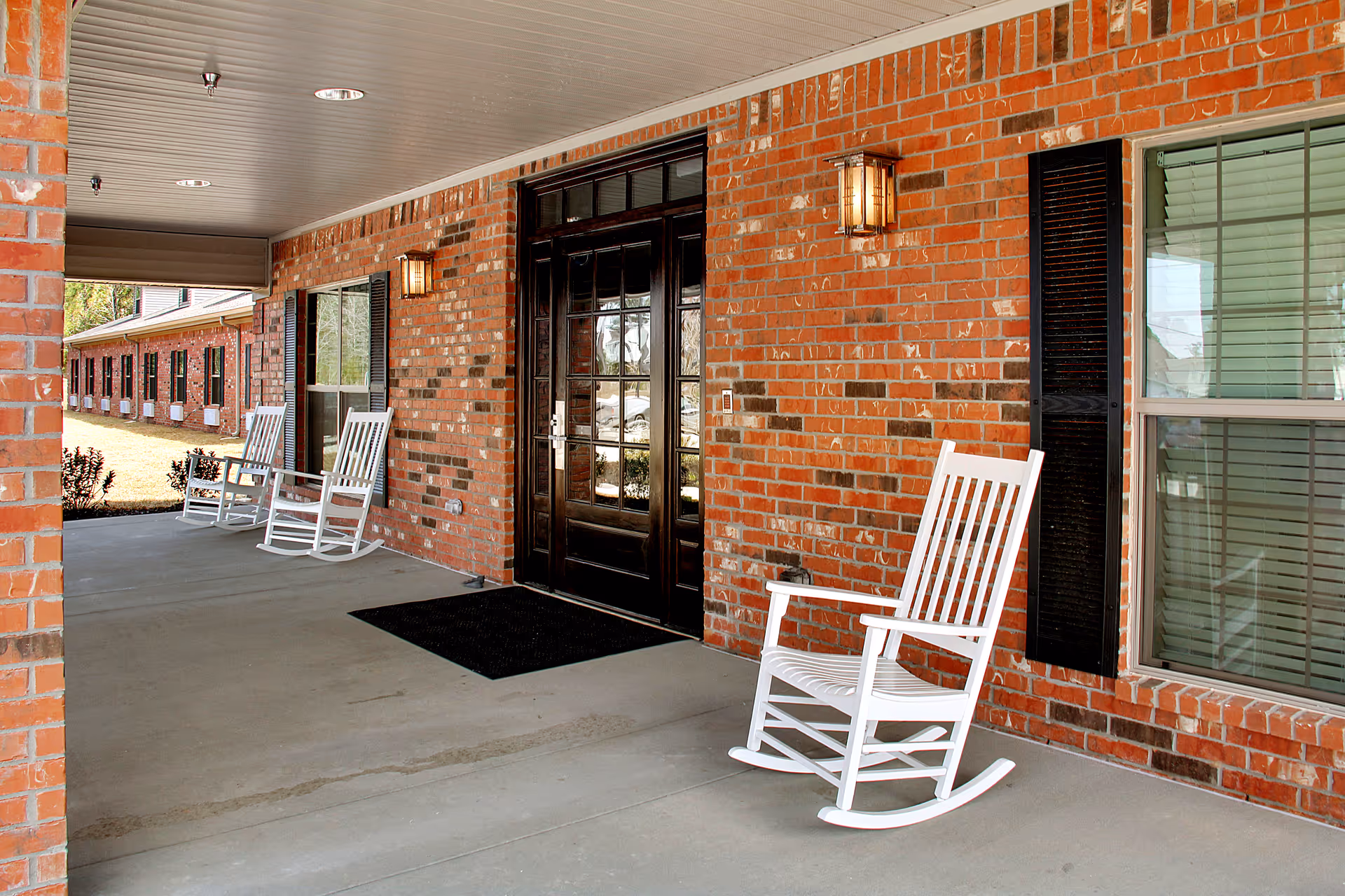 Covered porch area of a brick building with three white rocking chairs placed along the wall near windows and a dark wooden door with glass panels. The porch has a concrete floor and ceiling lights, with outdoor wall lanterns mounted on the brick wall.