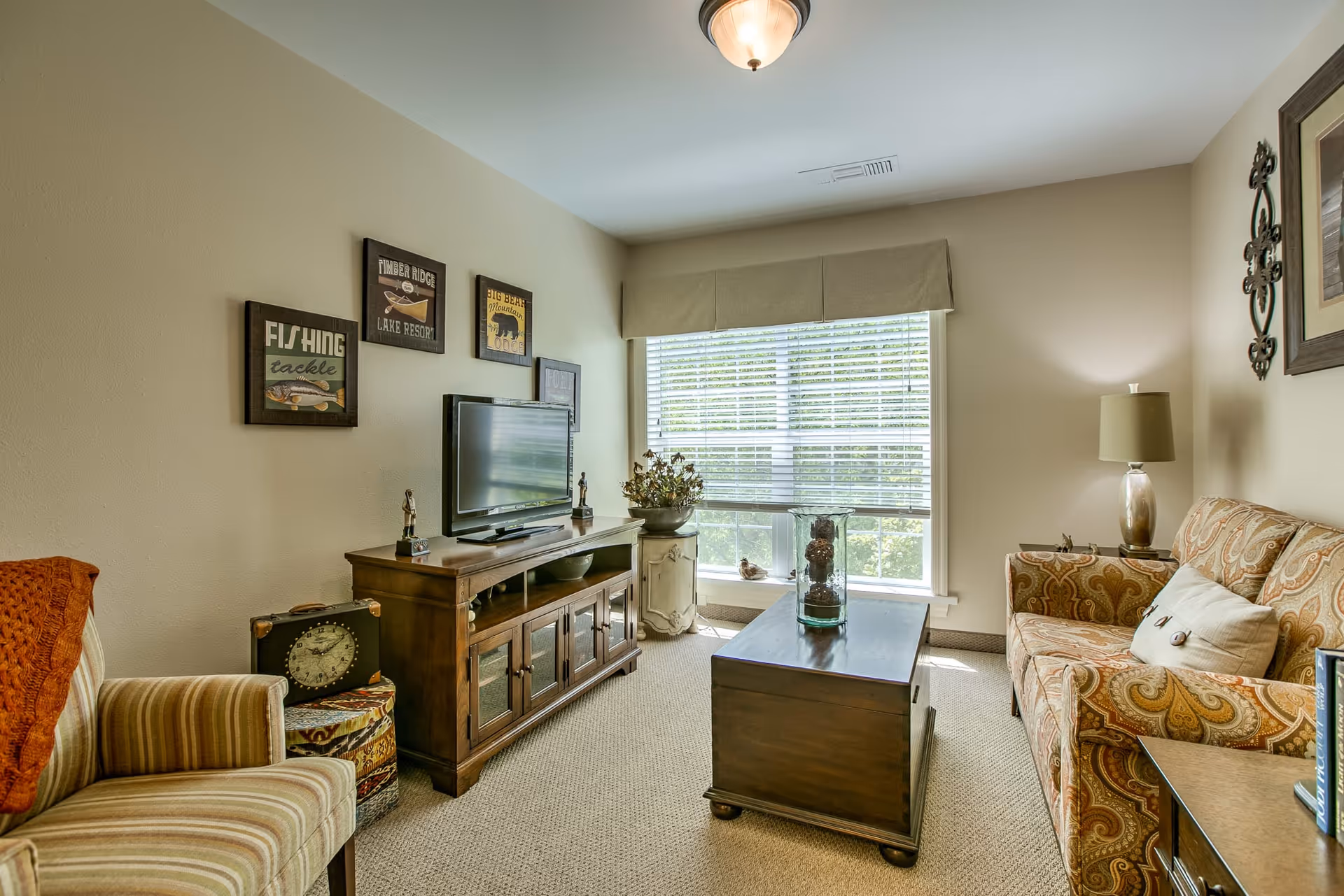A cozy living room with a patterned sofa, a striped armchair with an orange throw, a wooden coffee table, and a wooden TV stand with a flat-screen TV. The room has beige walls decorated with framed pictures and a window with blinds letting in natural light.