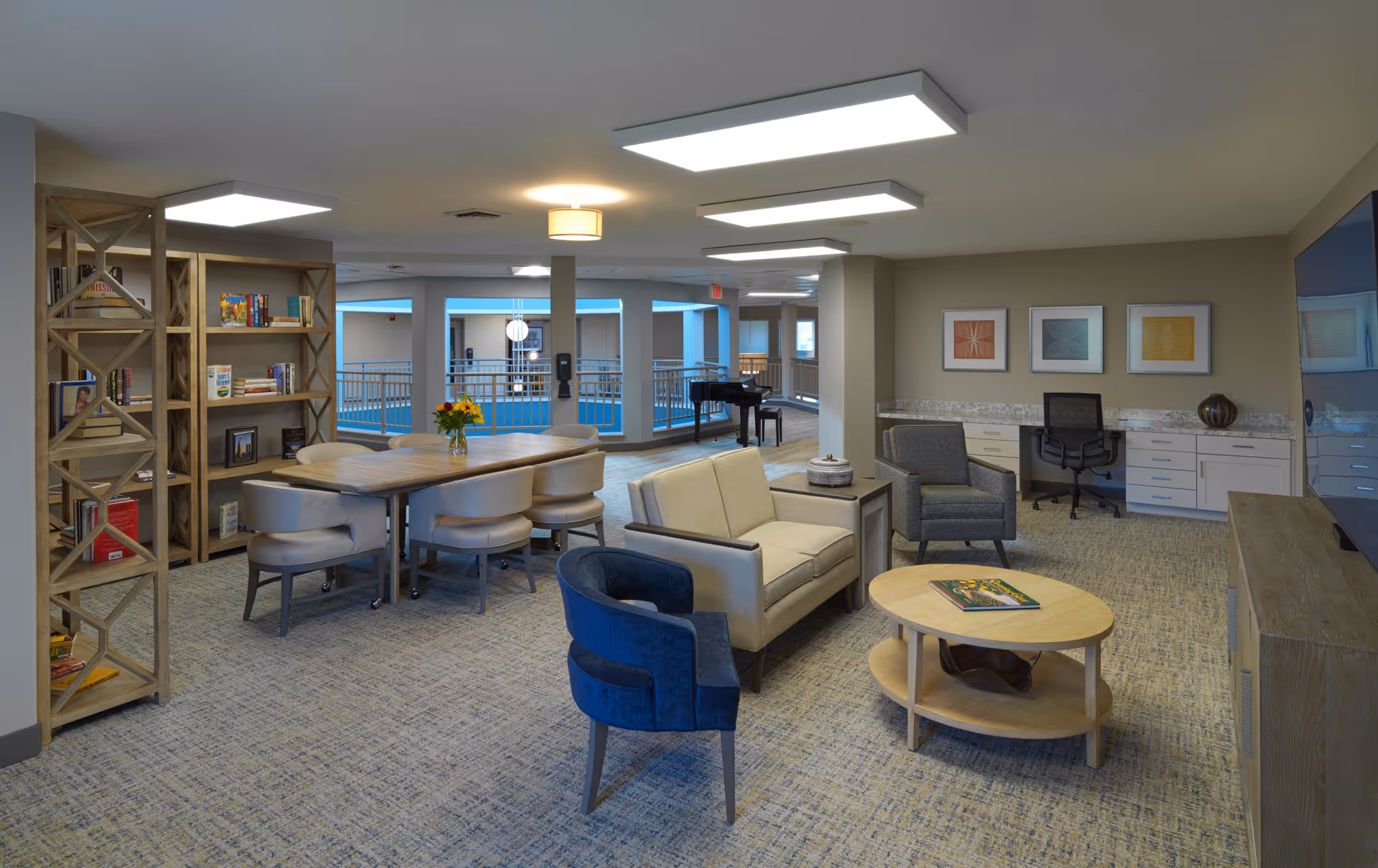 A bright lounge area with sofas, chairs, a round coffee table, dining table, bookshelf, and a desk overlooking an interior atrium.