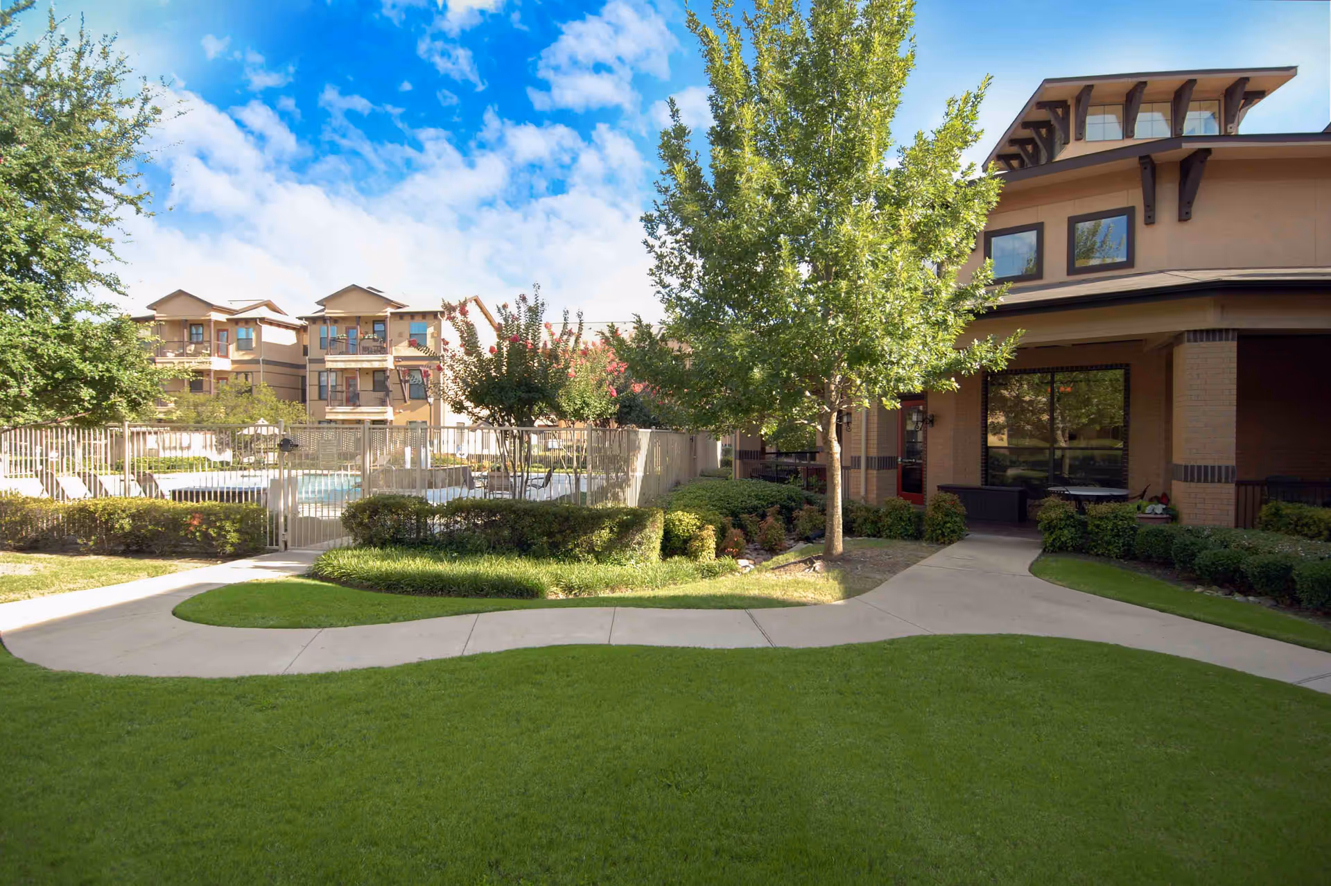 Outdoor view of a senior living facility with a well-maintained green lawn, paved walkways, trees, and bushes. There is a fenced swimming pool area with lounge chairs visible in the background, and a building with large windows and a covered porch on the right side under a partly cloudy blue sky.