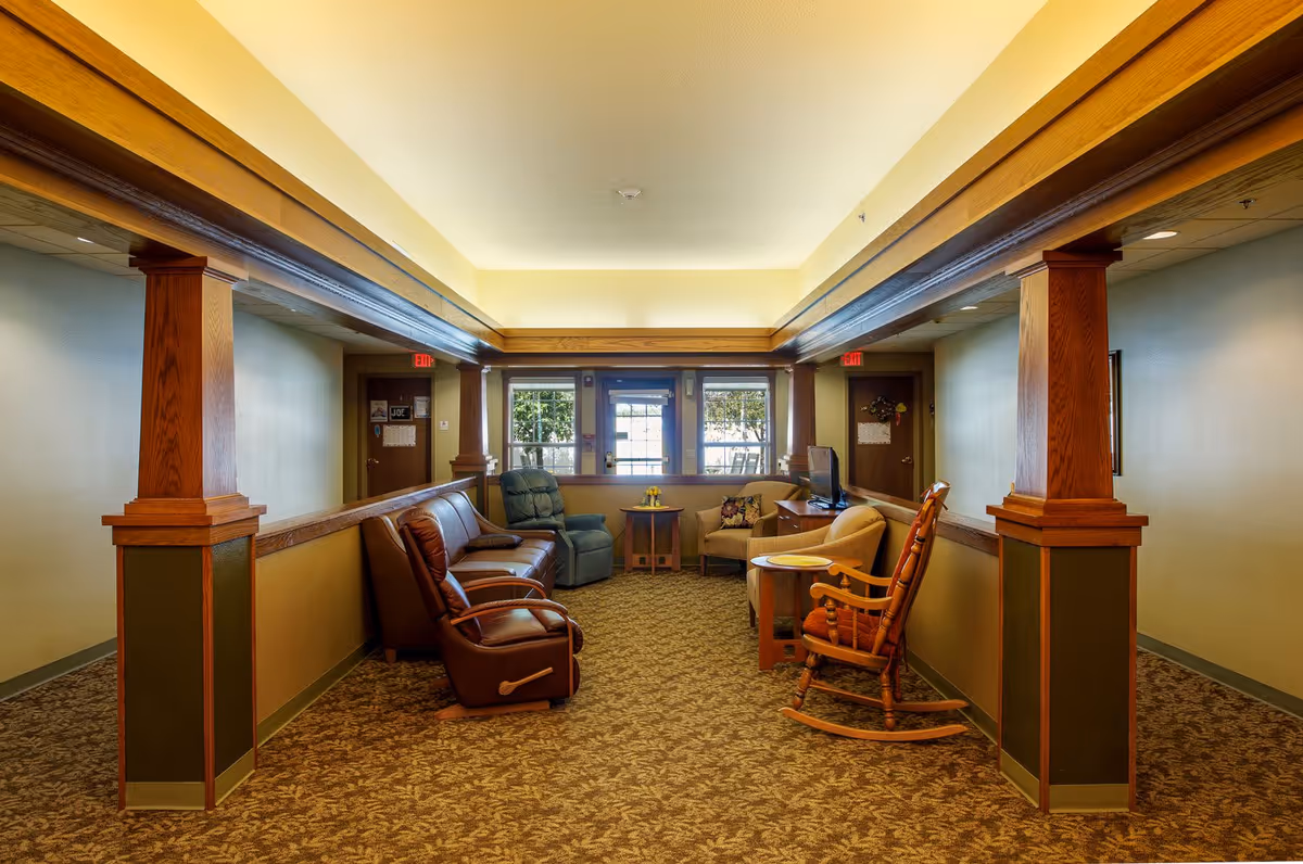 A cozy common area in an assisted living facility featuring a variety of seating options including a leather recliner, a blue armchair, a beige armchair, a rocking chair, and a leather sofa. The room has warm yellow walls, wooden pillars, and a carpeted floor with a leaf pattern. Large windows and a door at the far end let in natural light, and a small table with a vase of flowers is placed between the chairs.