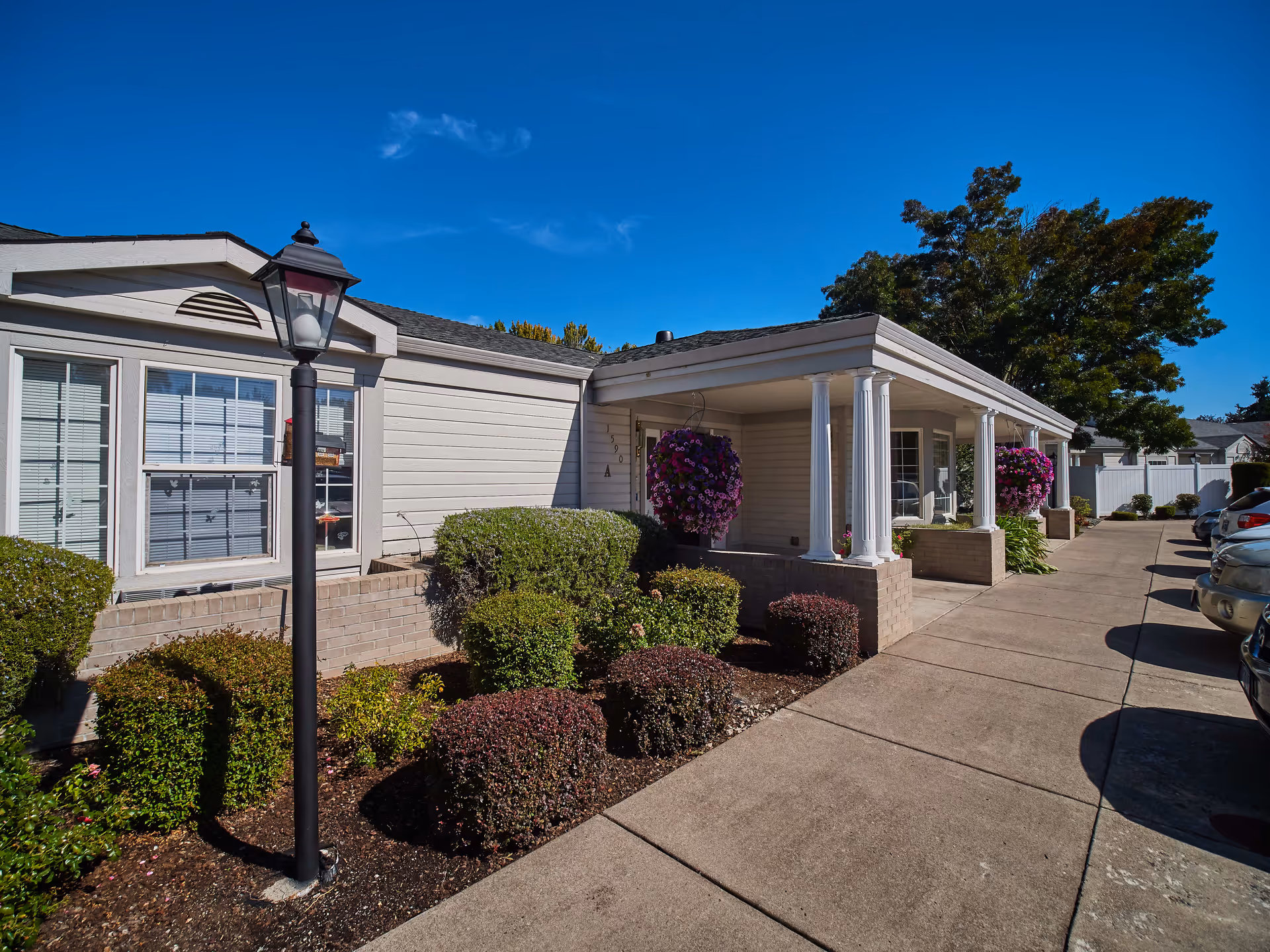 Exterior view of a single-story building with white siding and brick accents, featuring a covered porch supported by white columns. There are hanging flower baskets with purple flowers, neatly trimmed bushes, a black lamp post, and a concrete walkway leading to the entrance. Several parked cars are visible on the right side under a clear blue sky.