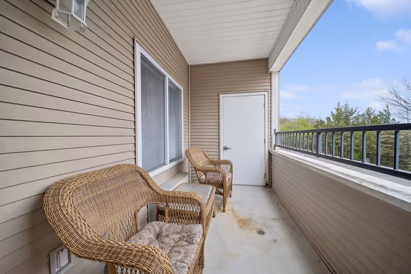A covered outdoor balcony area with beige siding walls, two wicker chairs with cushions, and a small wicker table. The balcony has a white ceiling, a white door at the end, and a black metal railing overlooking trees and a partly cloudy sky.