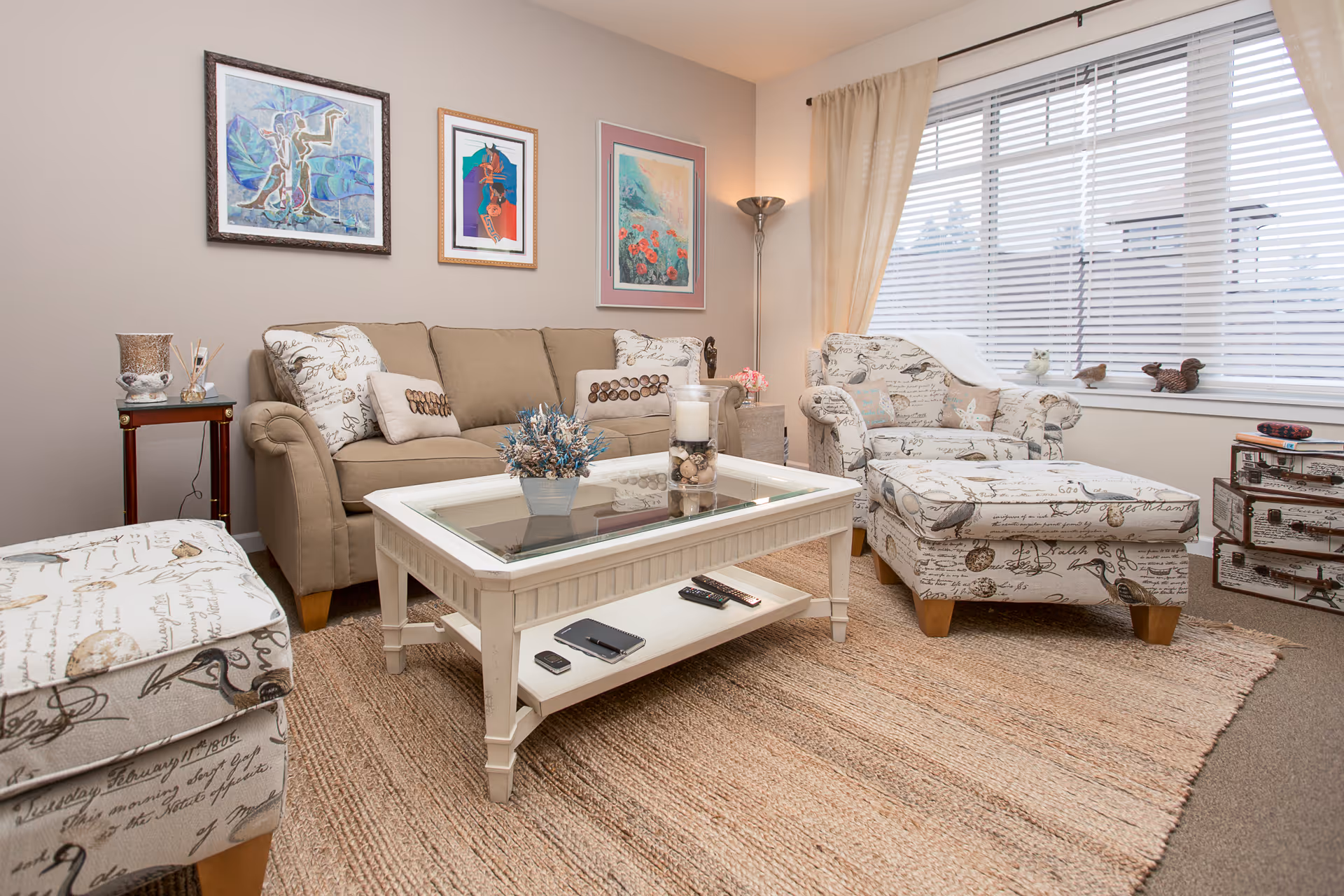 A cozy living room with a beige sofa adorned with decorative pillows, a matching armchair and ottoman with script and bird patterns, a white coffee table with glass top holding a small plant and candles, a side table with a lamp, and three framed artworks on the wall. The room has a large window with blinds and light curtains, and a woven area rug covering the floor.