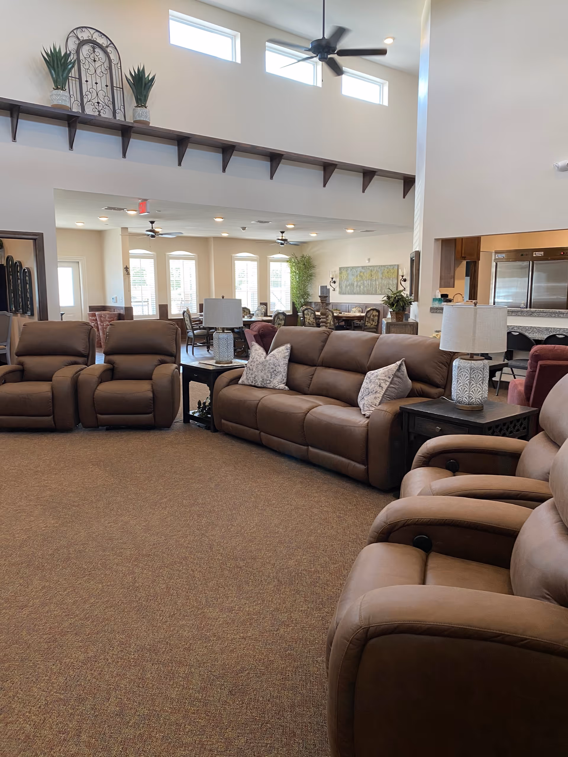 A spacious and well-lit living room area with several brown leather recliners and a matching sofa adorned with decorative pillows. The room features high ceilings with ceiling fans and small rectangular windows near the ceiling. There are side tables with lamps, and in the background, a dining area with tables and chairs is visible along with large windows letting in natural light. The walls are light-colored, and there are decorative plants and artwork adding to the cozy atmosphere.