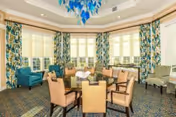 A bright and spacious living room area with large windows covered by floral curtains, featuring a round table surrounded by six beige upholstered chairs, two teal armchairs, and additional seating along the walls. The ceiling has a decorative blue chandelier.