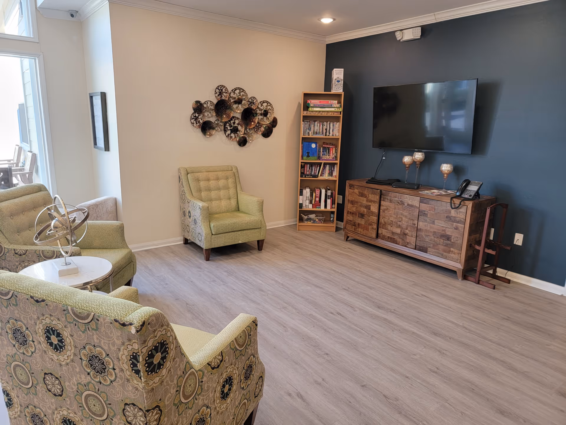 A cozy living room area with patterned and solid green armchairs arranged around a small round table with a decorative metal sculpture. A wooden bookshelf filled with books and games stands against a beige wall, and a flat-screen TV is mounted on a dark blue accent wall above a wooden cabinet. The room has light wood flooring and a large window letting in natural light.