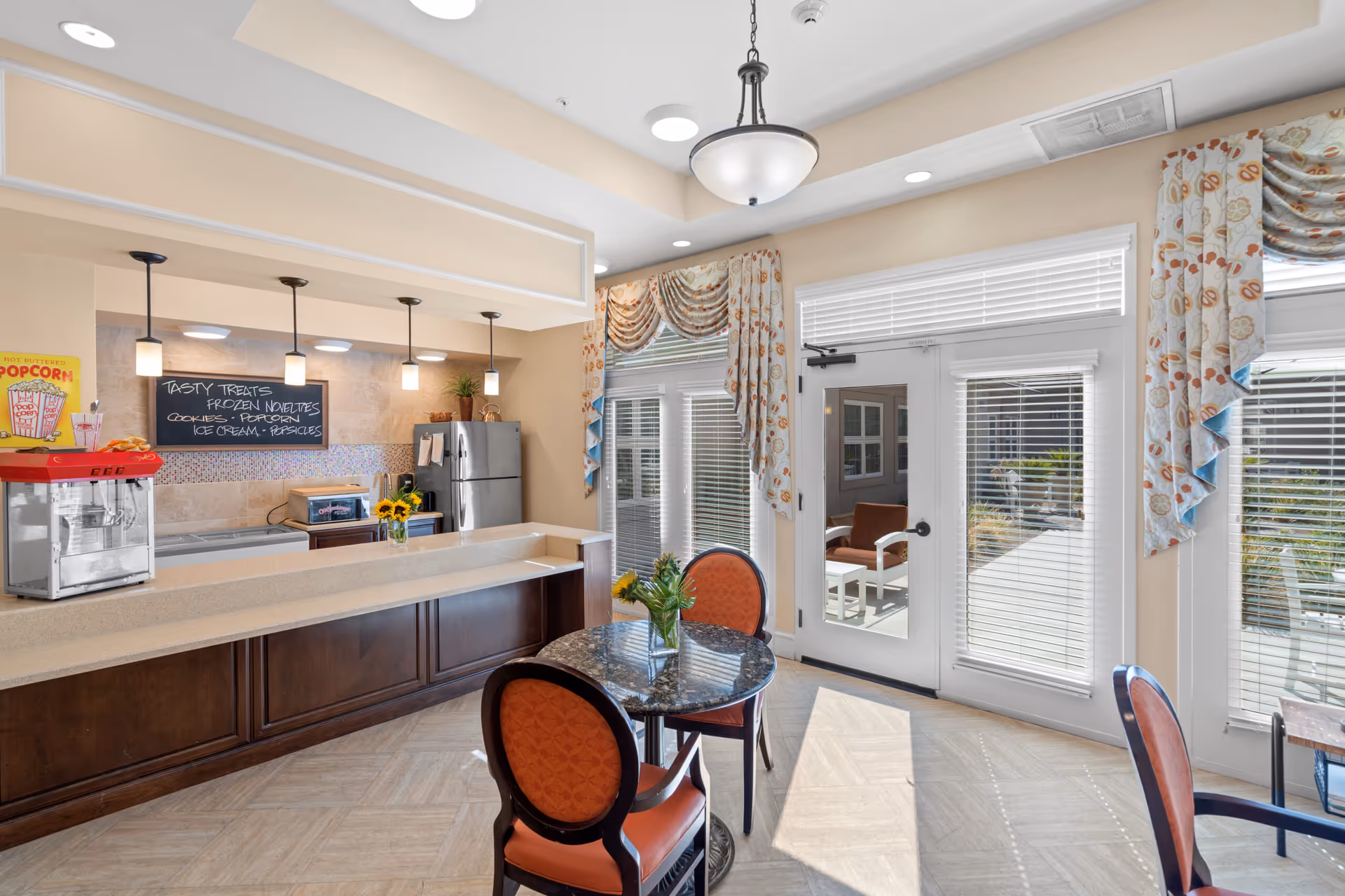 A bright and inviting snack area in a senior living facility featuring a countertop with a popcorn machine, a chalkboard menu listing tasty treats like frozen novelties, cookies, popcorn, ice cream, and popsicles. There is a small round table with two orange cushioned chairs and a vase with sunflowers. Large windows and glass doors with patterned curtains let in natural light and provide a view of an outdoor seating area.