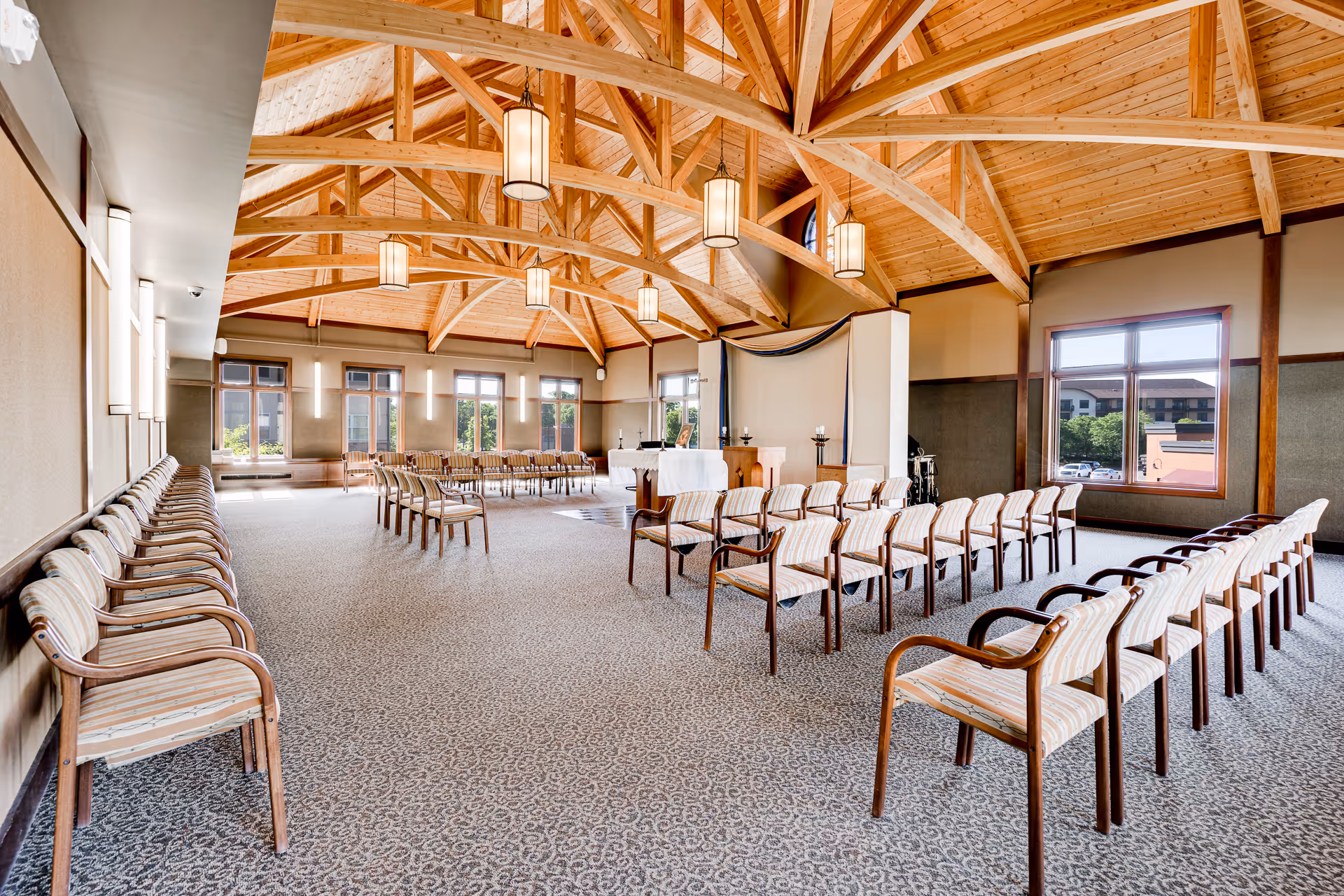 A spacious room with a high wooden vaulted ceiling featuring exposed beams and hanging pendant lights. The room is arranged with multiple rows of wooden chairs with striped cushions facing a small altar or podium area at the front. Large windows on the walls allow natural light to fill the space, and the carpet has a subtle patterned design.
