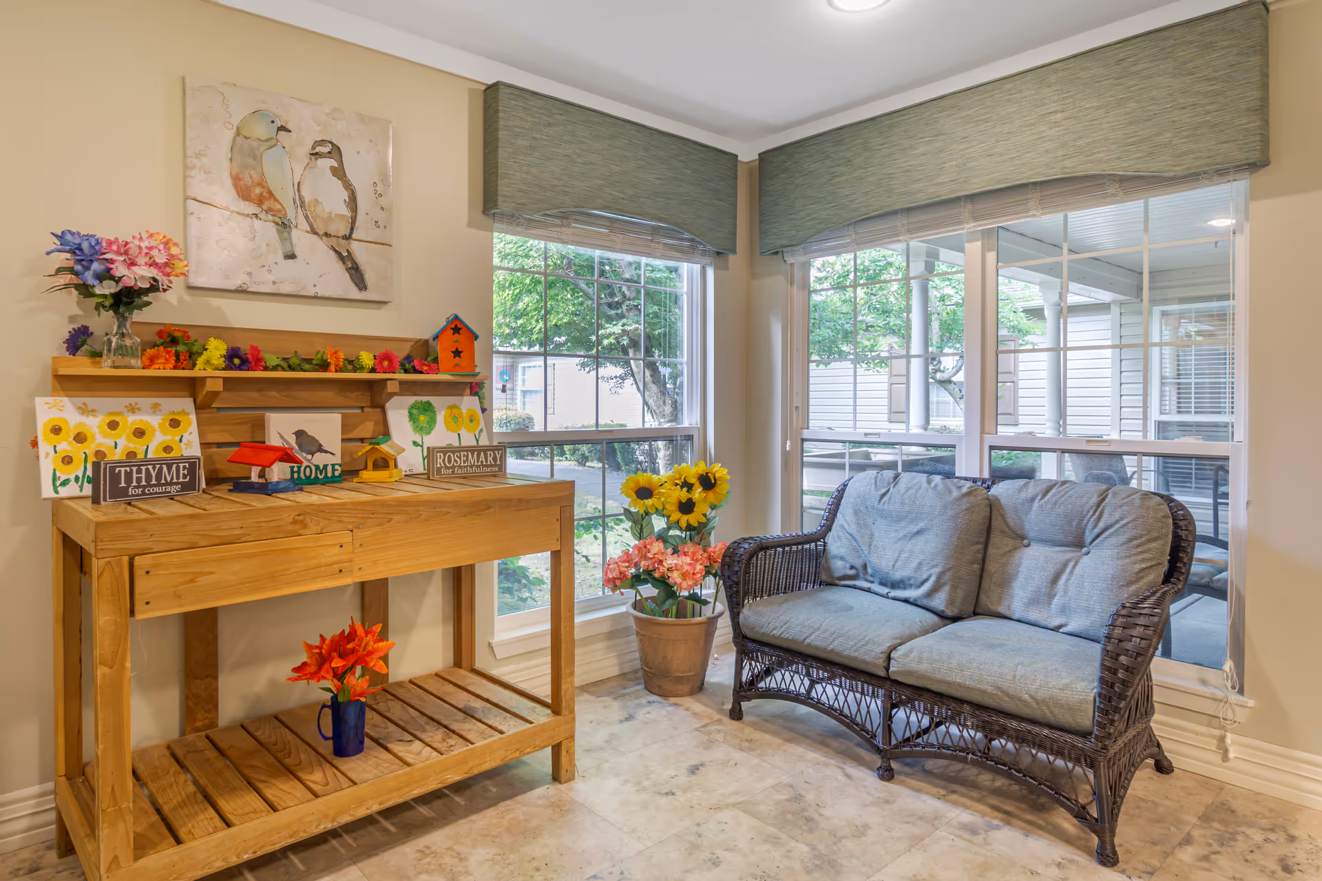 A cozy corner in a senior living facility with a wooden table decorated with colorful flower paintings, small birdhouses, and signs labeled 'THYME for courage', 'HOME', and 'ROSEMARY for faithfulness'. Next to the table is a wicker loveseat with blue cushions, positioned in front of large windows with green valances, showing a view of trees and the exterior of the building.
