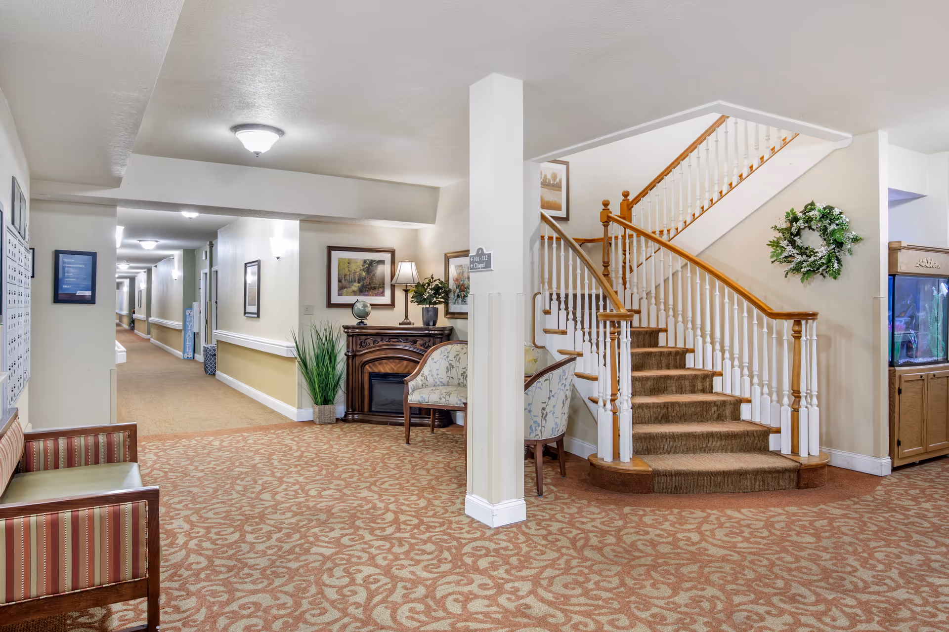 Interior view of a senior living facility hallway with patterned carpet, a staircase with wooden handrails, a small seating area with chairs, a decorative wreath on the wall, framed artwork, a small table with a lamp and plants, and an aquarium cabinet.