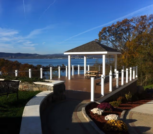 Outdoor patio area with a wooden deck and a white gazebo overlooking a large body of water. The area is surrounded by a white railing and has benches and picnic tables. There are landscaped flower beds with colorful flowers and trees nearby under a clear blue sky.