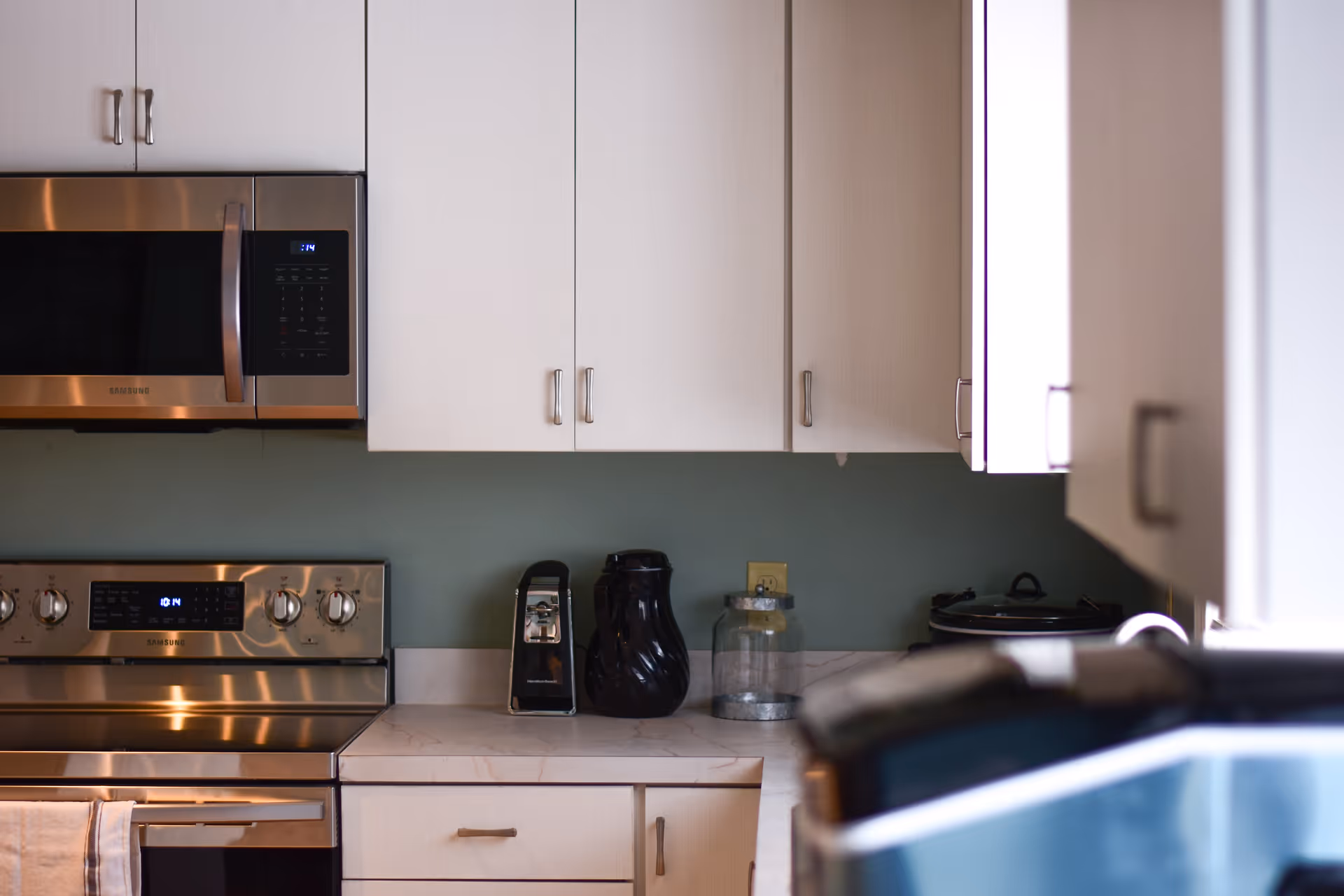 A modern kitchen with white cabinets, a stainless steel microwave and stove, a black coffee maker, a black pitcher, and a glass jar on the countertop. The wall behind the counter is painted green.