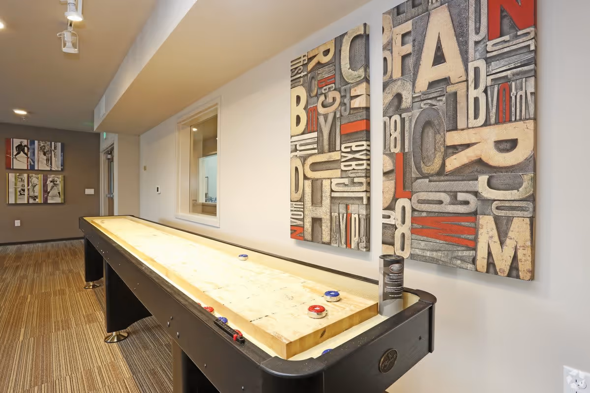 Indoor game room featuring a long shuffleboard table and large typographic wall art on a neutral wall.