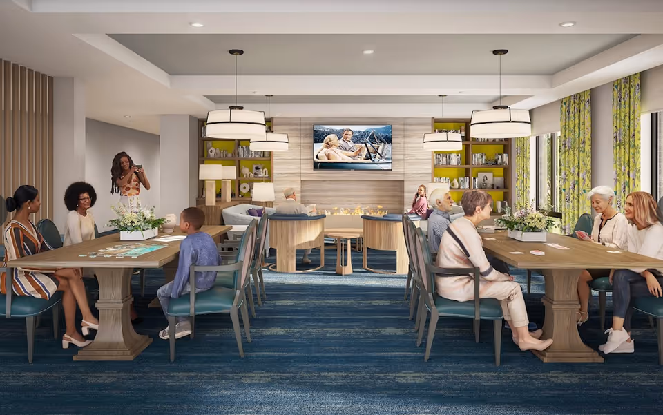 A spacious and well-lit common area in a senior living facility with two long tables where people are engaged in activities like playing cards and puzzles. In the background, there is a cozy seating area with armchairs around a modern fireplace and a wall-mounted TV. The room has large windows with floral curtains, bookshelves, and pendant lights hanging from the ceiling.