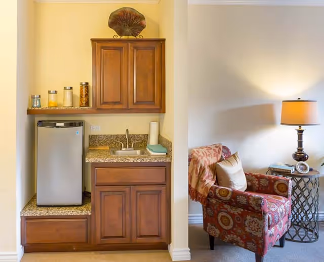 A small kitchenette area with wooden cabinets, a granite countertop, a mini refrigerator, a sink, and some jars on the shelf. Next to the kitchenette is a cozy sitting area with a patterned armchair, a side table with books, and a lit table lamp against a plain wall.