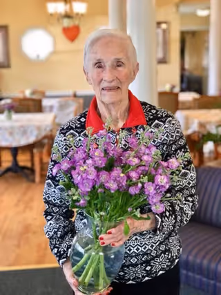 An elderly woman with short white hair wearing a black and white patterned sweater with a red collar is holding a clear vase filled with purple flowers. She is standing in a warmly lit room with tables covered in floral tablecloths and chairs in the background.