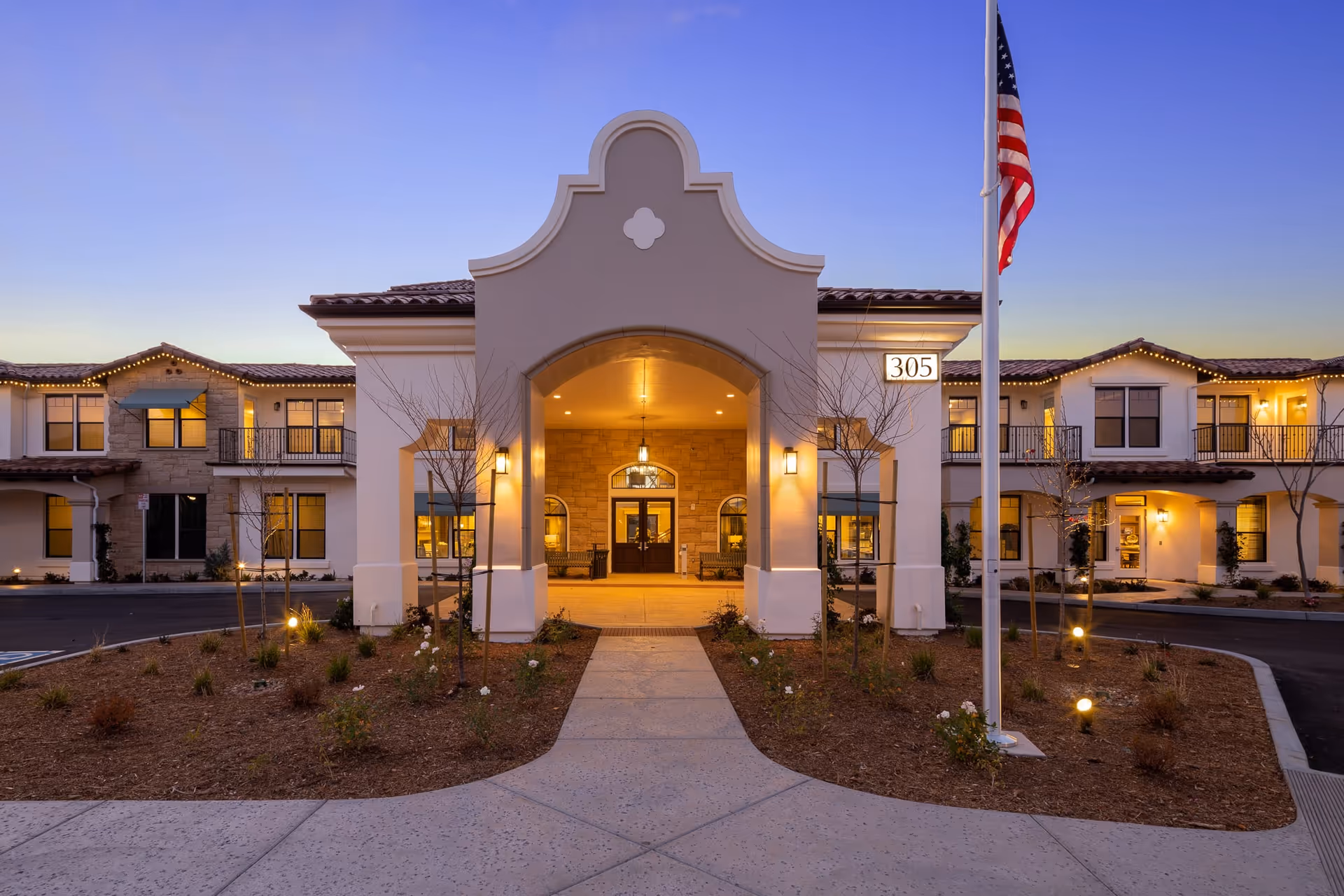 Front entrance of a two-story residential building at dusk with an arched covered entry, lit windows, landscaping, and an American flag.