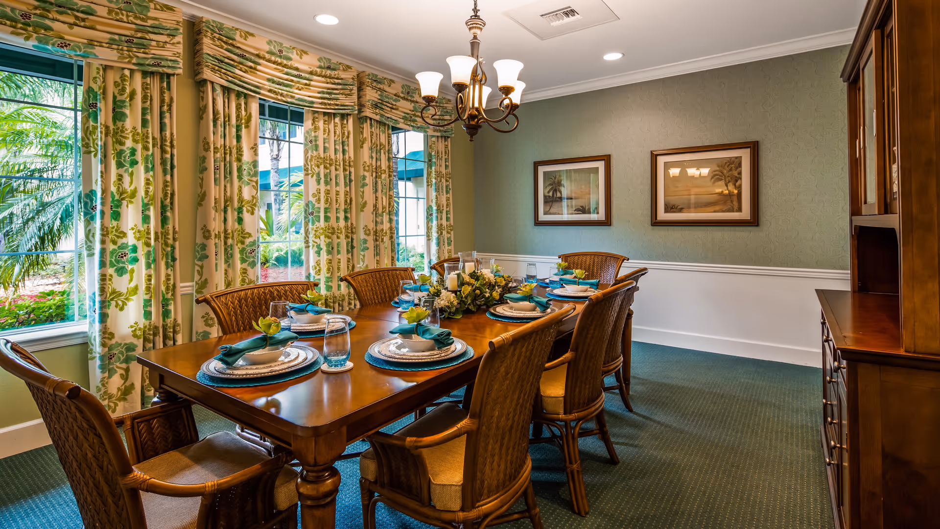 A formal dining room with a wooden dining table set for six people. The table is decorated with plates, glasses, napkins, and a floral centerpiece. The room has large windows with floral curtains, green carpet, and two framed pictures on the wall. A chandelier hangs above the table.