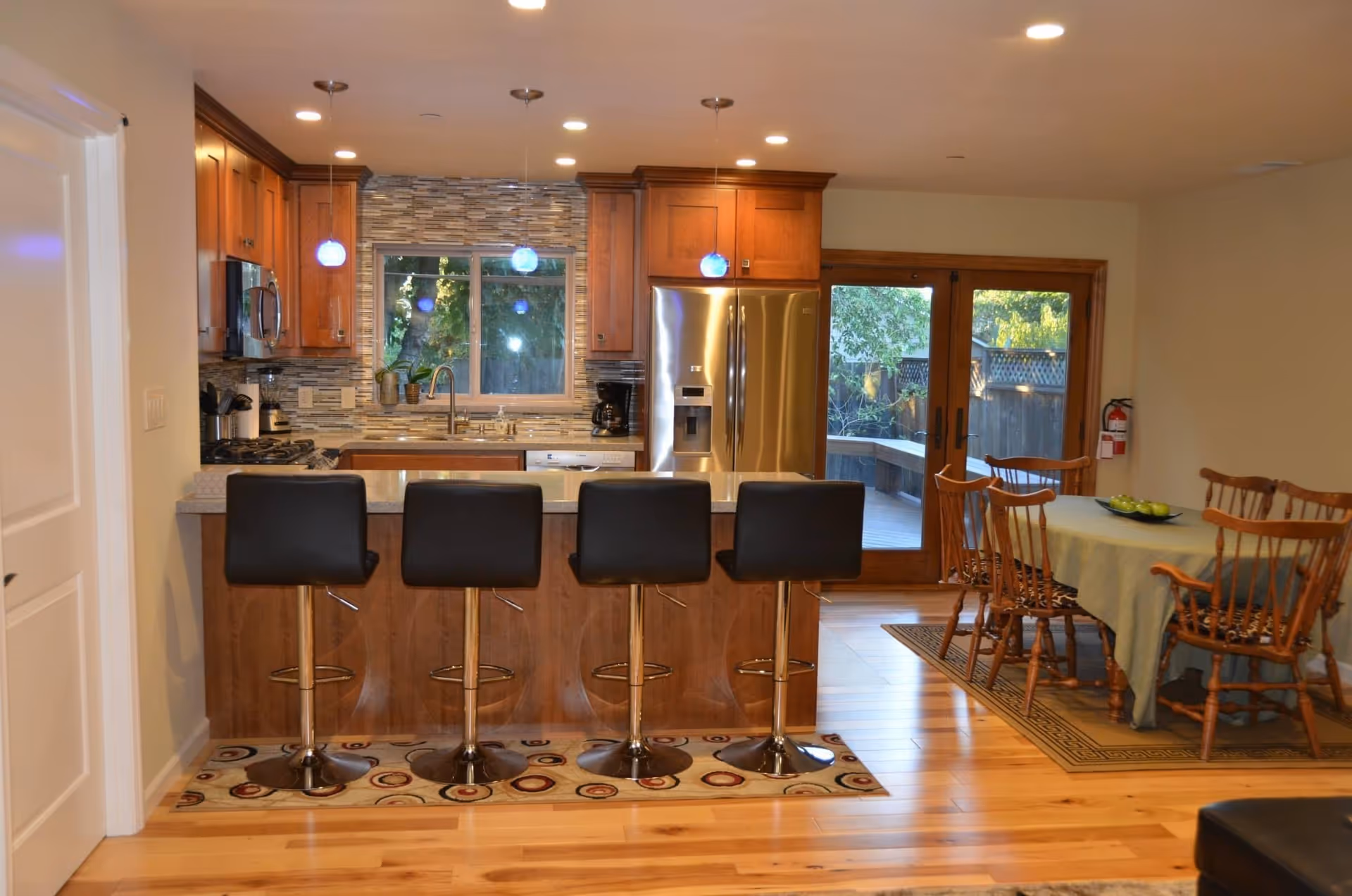 Modern kitchen and dining area with wooden cabinets, stainless steel refrigerator, and a kitchen island with four black bar stools. The dining table is covered with a green tablecloth and surrounded by wooden chairs. Large glass doors lead to an outdoor area with greenery visible outside.