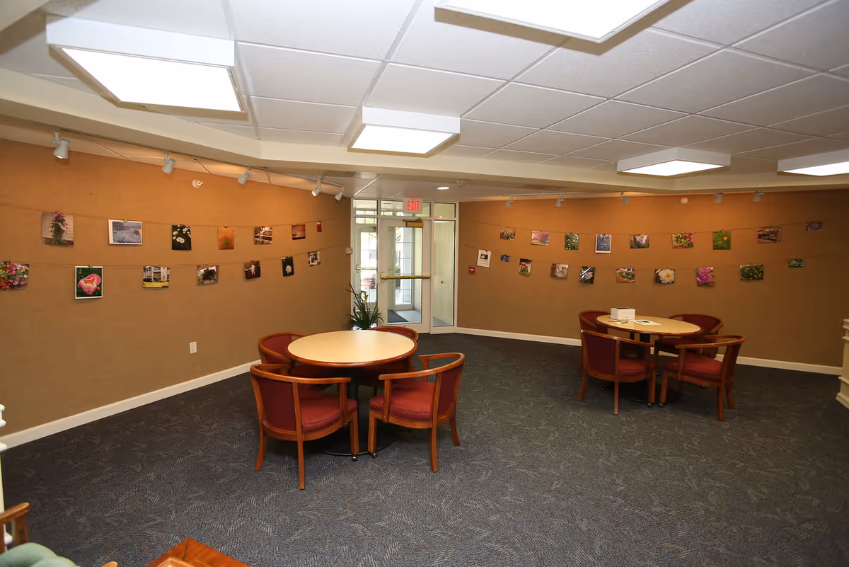 A common area in Melrose Meadows Retirement Community featuring two round tables each surrounded by four wooden chairs with red cushions. The walls are decorated with strings of photographs, mostly of flowers and nature scenes. The room has a carpeted floor, beige walls, and a ceiling with recessed lighting. A glass door with an exit sign above it is visible at the back of the room.