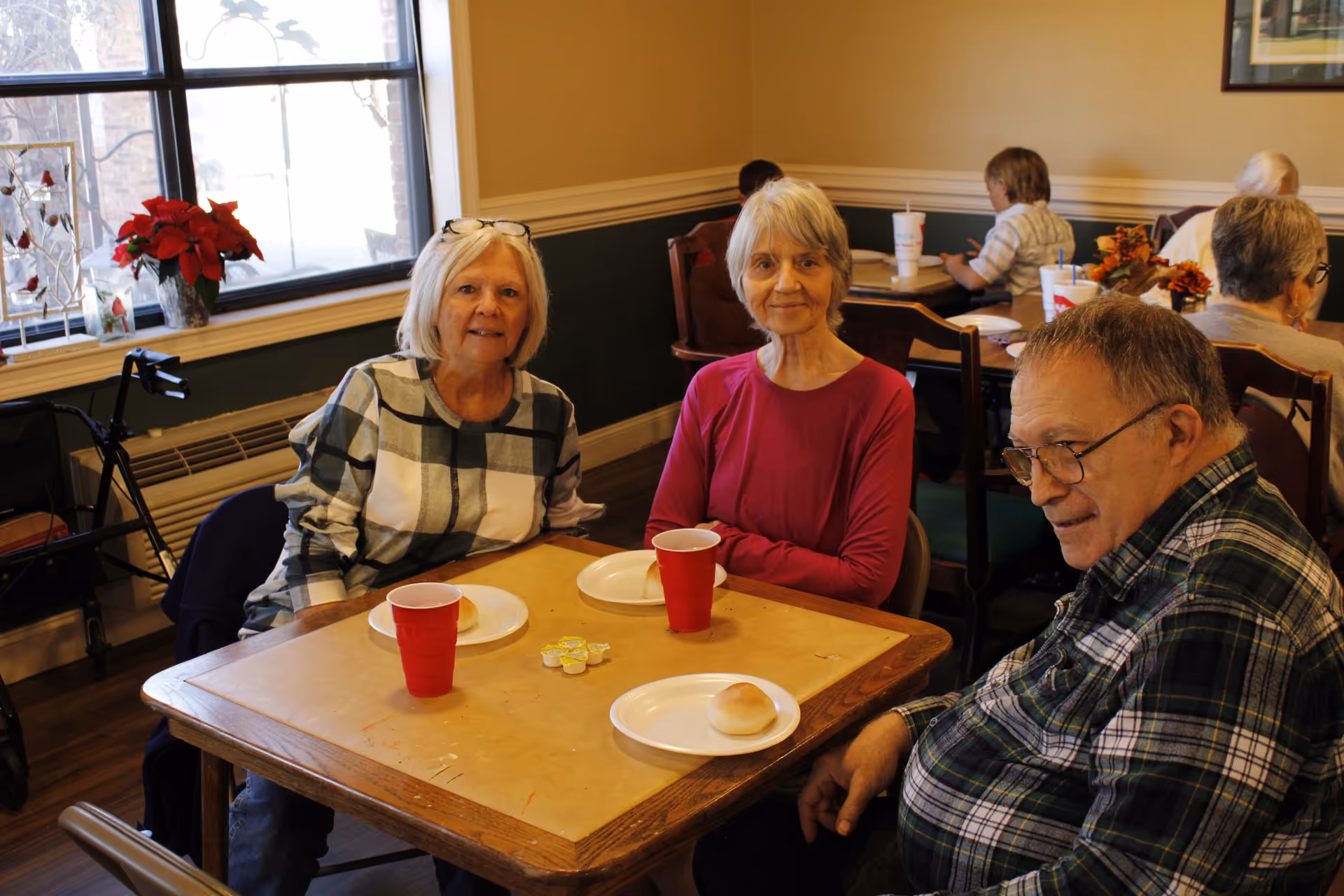 Three elderly people sitting around a wooden table in a dining area, each with a plate holding a bread roll and a red cup. The room has large windows with a poinsettia plant on the windowsill and other people seated at tables in the background.
