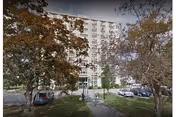 Exterior view of a multi-story residential building partially obscured by trees with autumn foliage. There is a pathway leading to the entrance and several parked cars in front of the building.