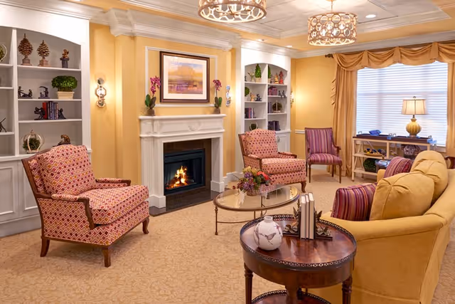 A cozy living room with yellow walls and carpet, featuring a white fireplace with a fire burning inside. Two patterned armchairs and a yellow sofa surround a glass coffee table with a flower arrangement. Built-in white shelves with decorative items flank the fireplace. A window with yellow curtains and a table lamp is in the background. Two ceiling light fixtures hang from the ceiling.