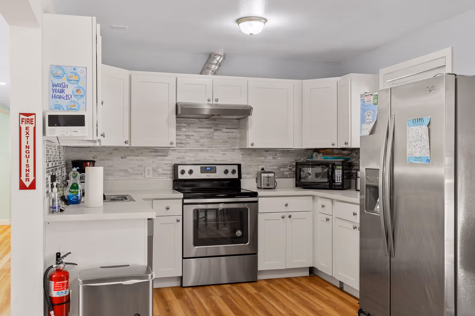 A clean, modern kitchen with white cabinets, stainless steel appliances including a stove, microwave, and refrigerator. The kitchen has a light gray tiled backsplash and wood flooring. A fire extinguisher is mounted on the wall next to a sign that says 'FIRE EXTINGUISHER' and a poster reminding to wash hands. There is a paper towel roll and dish soap on the counter.