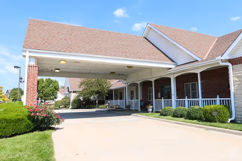 Covered entrance/porte-cochere of a brick senior living building with a driveway, porch, and landscaping under a clear blue sky.
