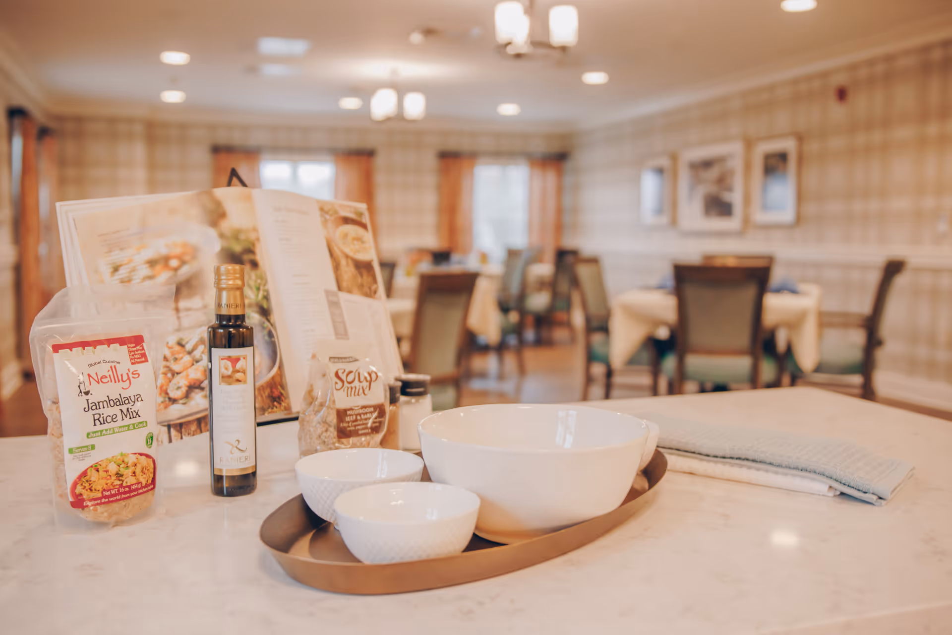 Countertop display of bowls, a bottle of oil and packaged mixes in front of a softly lit dining room with tables and chairs.