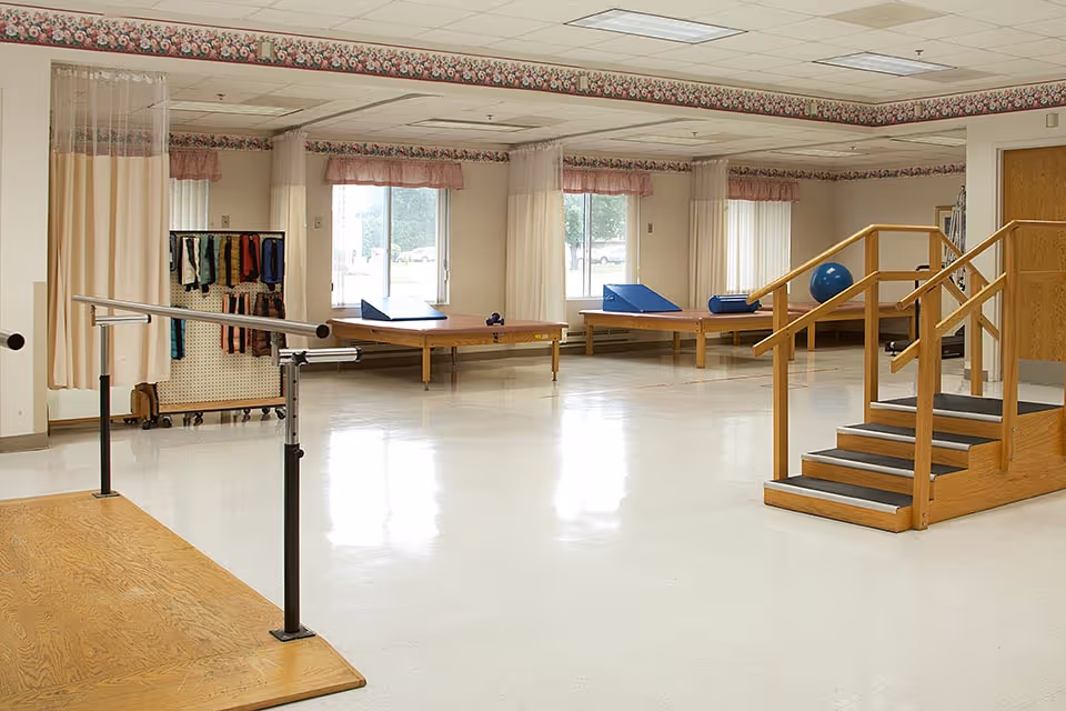 A spacious physical therapy or rehabilitation room with parallel bars for walking practice, wooden stairs with handrails for step training, padded therapy tables, and various exercise equipment. The room has large windows with sheer curtains and floral wallpaper border near the ceiling.