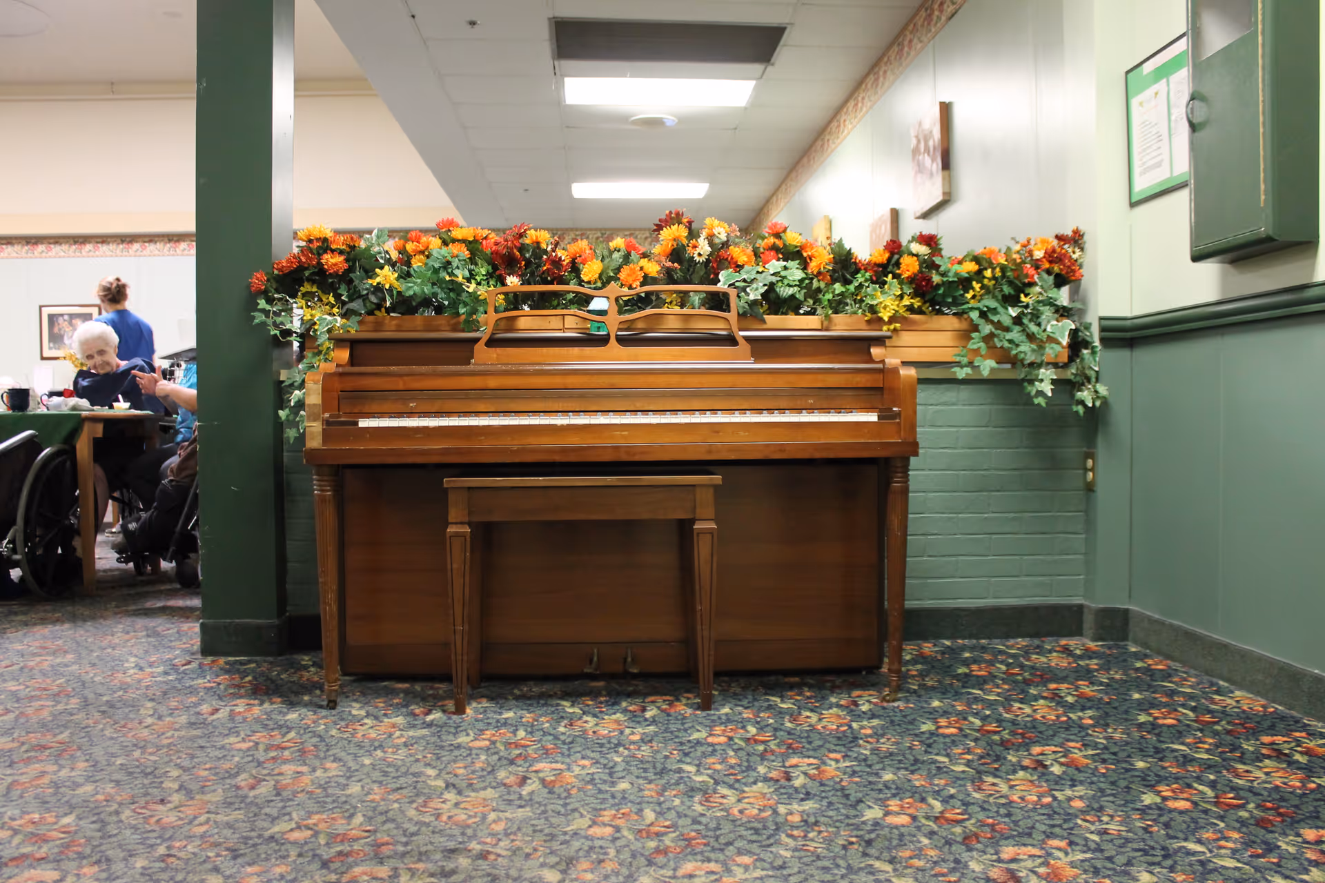 A wooden upright piano with a matching bench in front of it, decorated with a planter box filled with colorful artificial flowers on top. The piano is placed in a hallway with green walls and floral carpet. In the background, elderly people are seated at tables, with one person in a wheelchair and a caregiver standing nearby.
