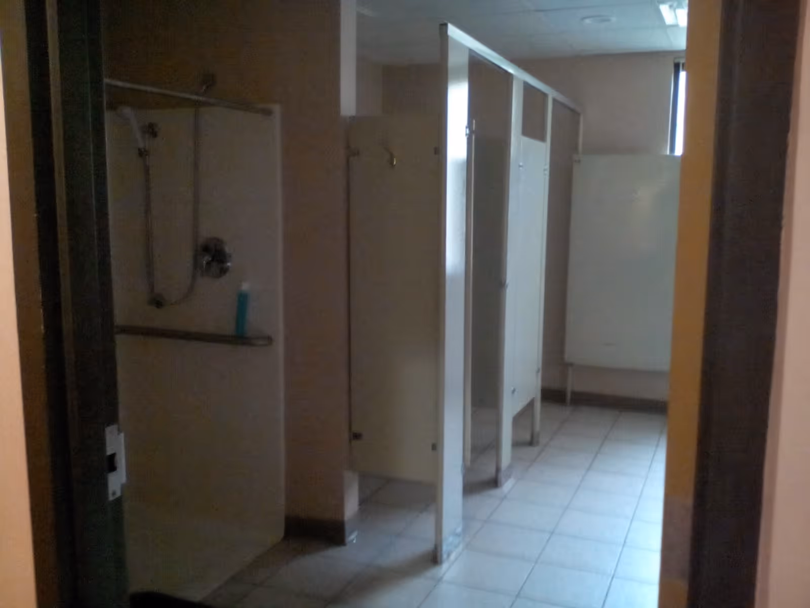 A dimly lit communal bathroom with multiple shower stalls and toilet cubicles. The floor is tiled, and there is a window at the far end letting in some natural light.
