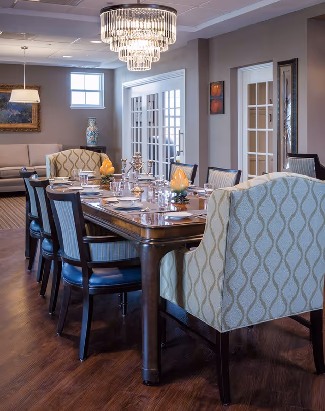 A well-lit dining room with a long wooden table set for a meal, featuring plates, bowls, glasses, and decorative candle holders. The table is surrounded by upholstered chairs, including two patterned armchairs at the ends. A crystal chandelier hangs above the table, and in the background, there is a sofa, a framed painting, a decorative vase, and French doors leading to another room.