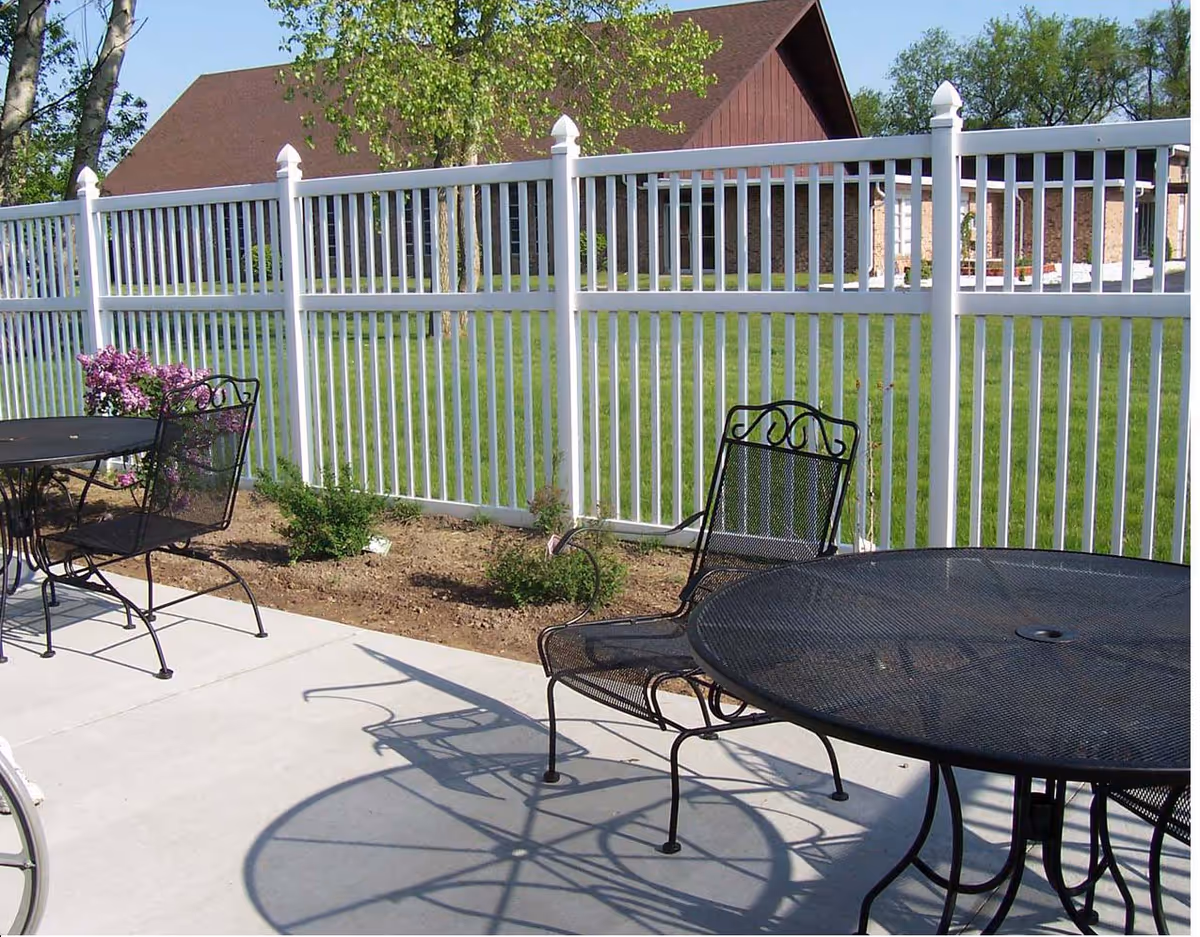 Outdoor patio area with black metal tables and chairs on a concrete surface, surrounded by a white picket fence. Green grass and a building with a brown roof are visible in the background.