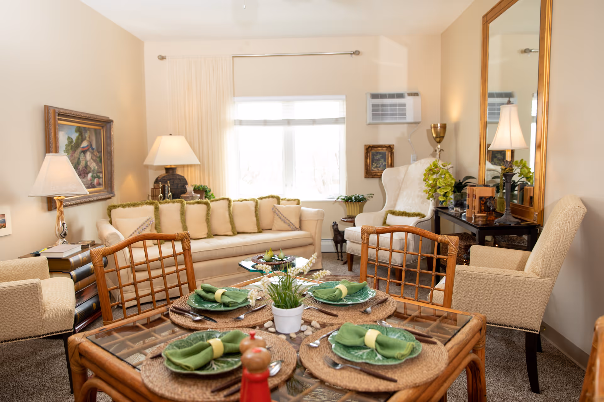 A bright living room with a dining table set for four in the foreground and a sofa and armchairs by a window.