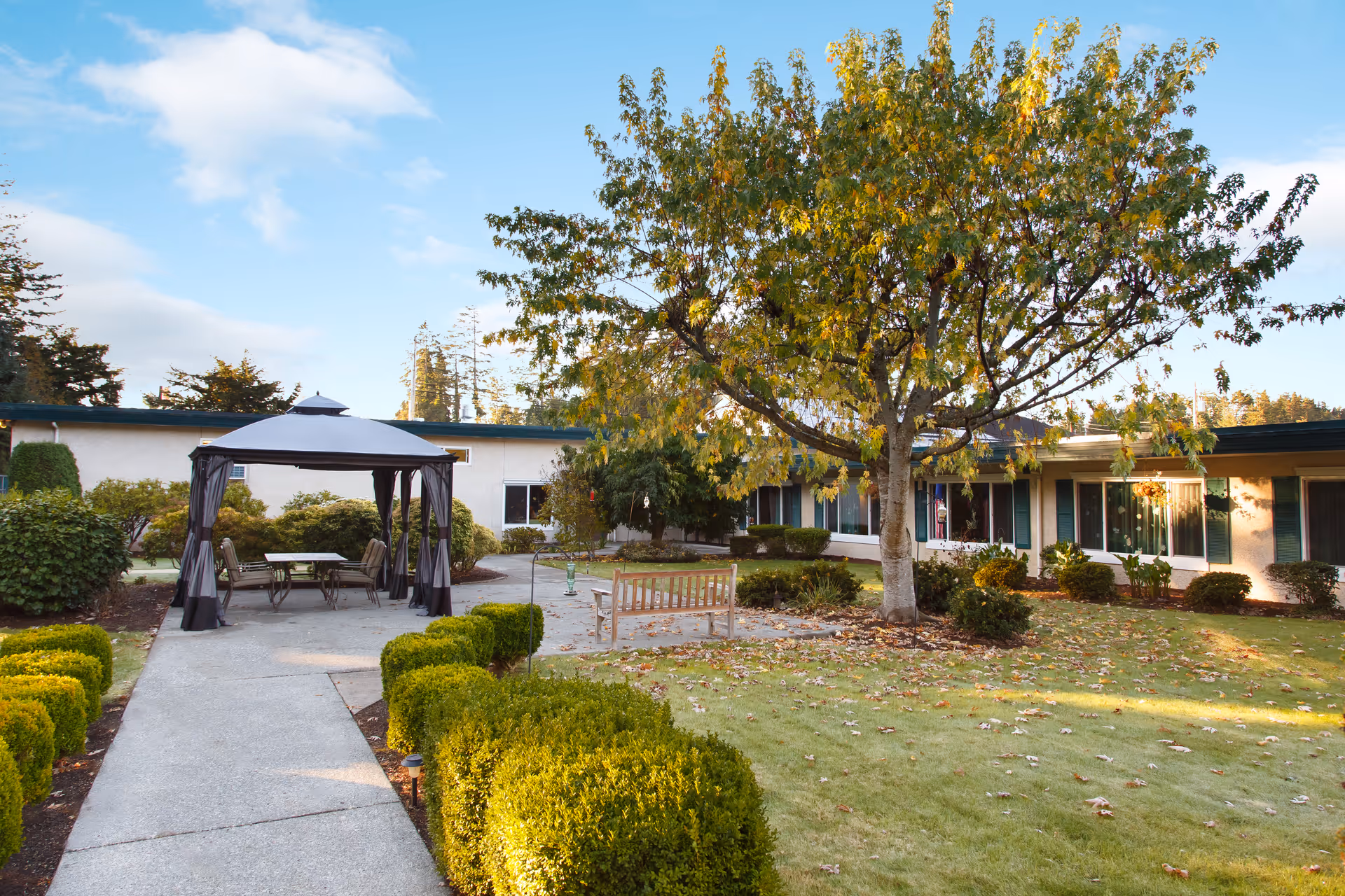 Outdoor garden area at Life Care Center of Coos Bay with a paved walkway, a wooden bench, a gazebo with a table and chairs underneath, surrounded by green bushes and trees with some autumn leaves on the ground.