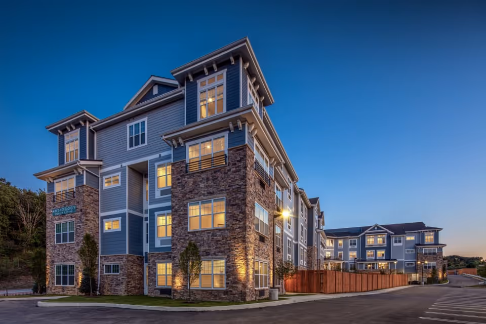 Exterior view of a modern multi-story senior living facility building at dusk with lights on inside. The building features a combination of stone and blue siding with multiple windows and a clear sky in the background.