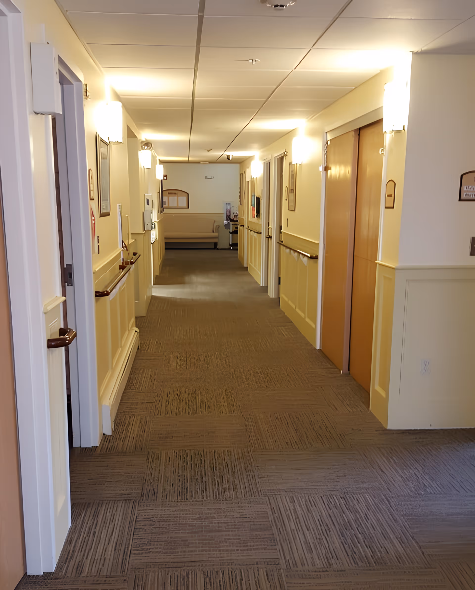 A well-lit hallway in a senior living facility with beige walls, carpeted floor, handrails on both sides, several doors, and a bench at the far end.