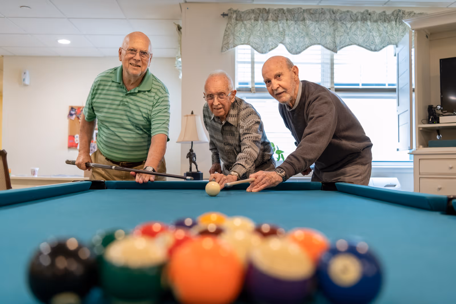 Three elderly men playing pool indoors, leaning over a pool table and aiming at the cue ball. The room has a window with a patterned valance, a lamp, and some furniture in the background.