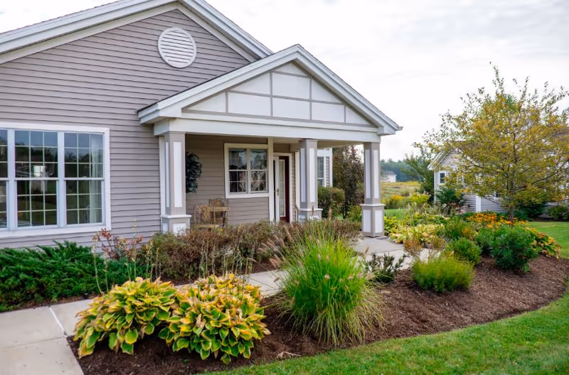 Front exterior view of a single-story building with gray siding, white trim, and a covered porch. The porch has two white columns and a small seating area with chairs. There is a well-maintained garden with various green plants and shrubs along the walkway leading to the entrance. A tree and more greenery are visible on the right side under a cloudy sky.