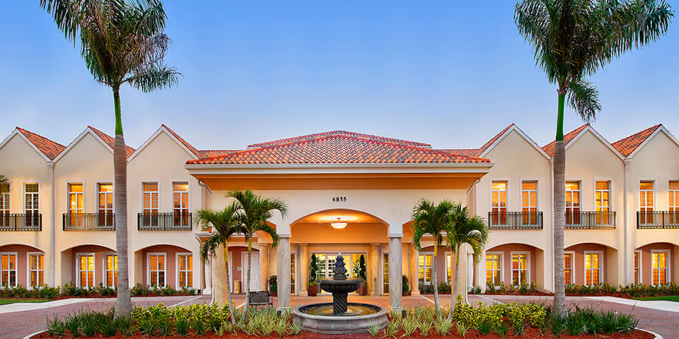 Mediterranean-style senior living building entrance with a central fountain, palm trees, and lit windows at dusk.