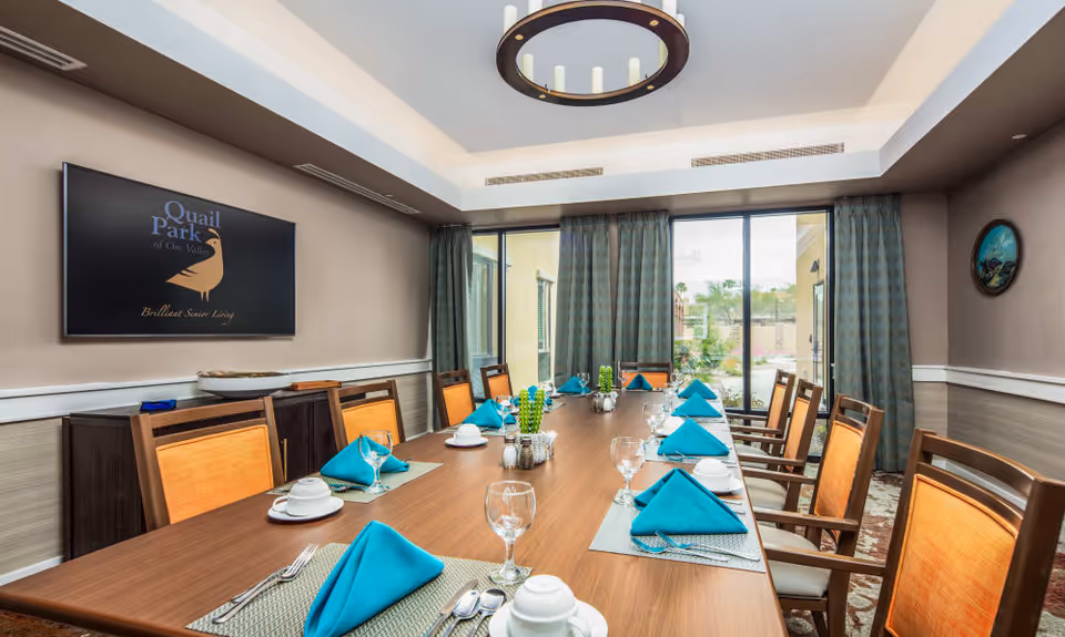 Bright dining room with a long wooden table set with turquoise napkins, place settings, chairs, and large windows with curtains.