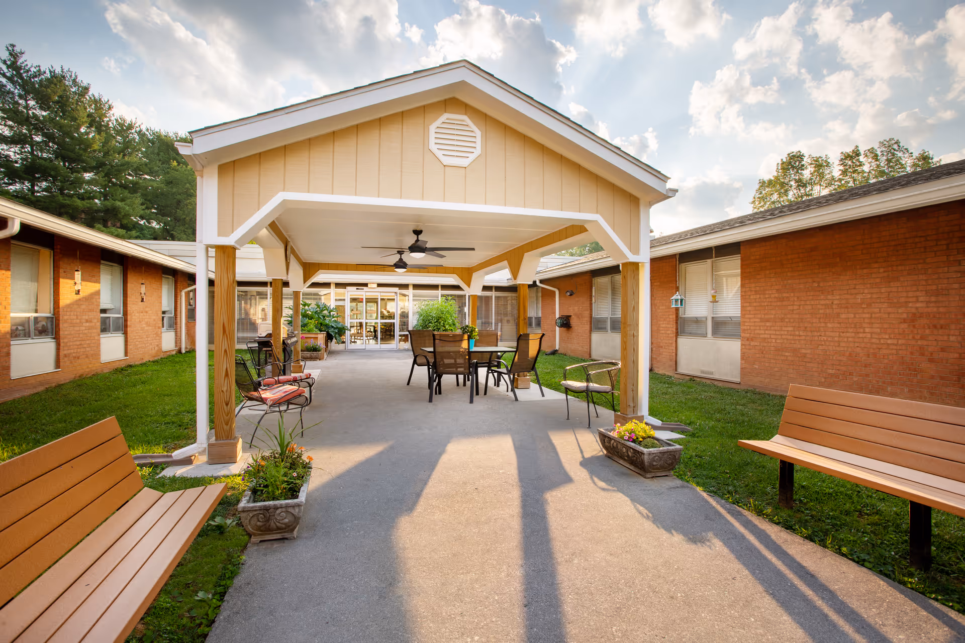 Covered outdoor courtyard with tables, chairs and benches between single-story brick buildings leading to a glass entrance.